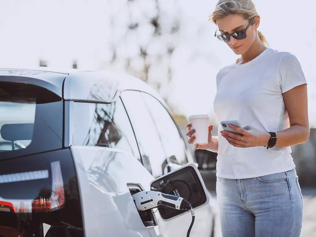 Eine junge Frau mit Smartphone und Kaffeebecher in der Hand steht neben ihrem elektrischen Auto das aufgeladet wird. E-Tankstelle. Umwelt.